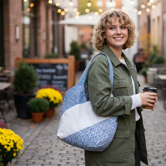 Smiling woman carrying the Blue Floral Patchwork Tote Bag over her shoulder while standing in a brick-lined outdoor market or cafe. The quilted bag features a blue floral pattern and a diagonal white stripe. The model is wearing a green utility jacket and holding a coffee, demonstrating the tote's casual, everyday wearability.